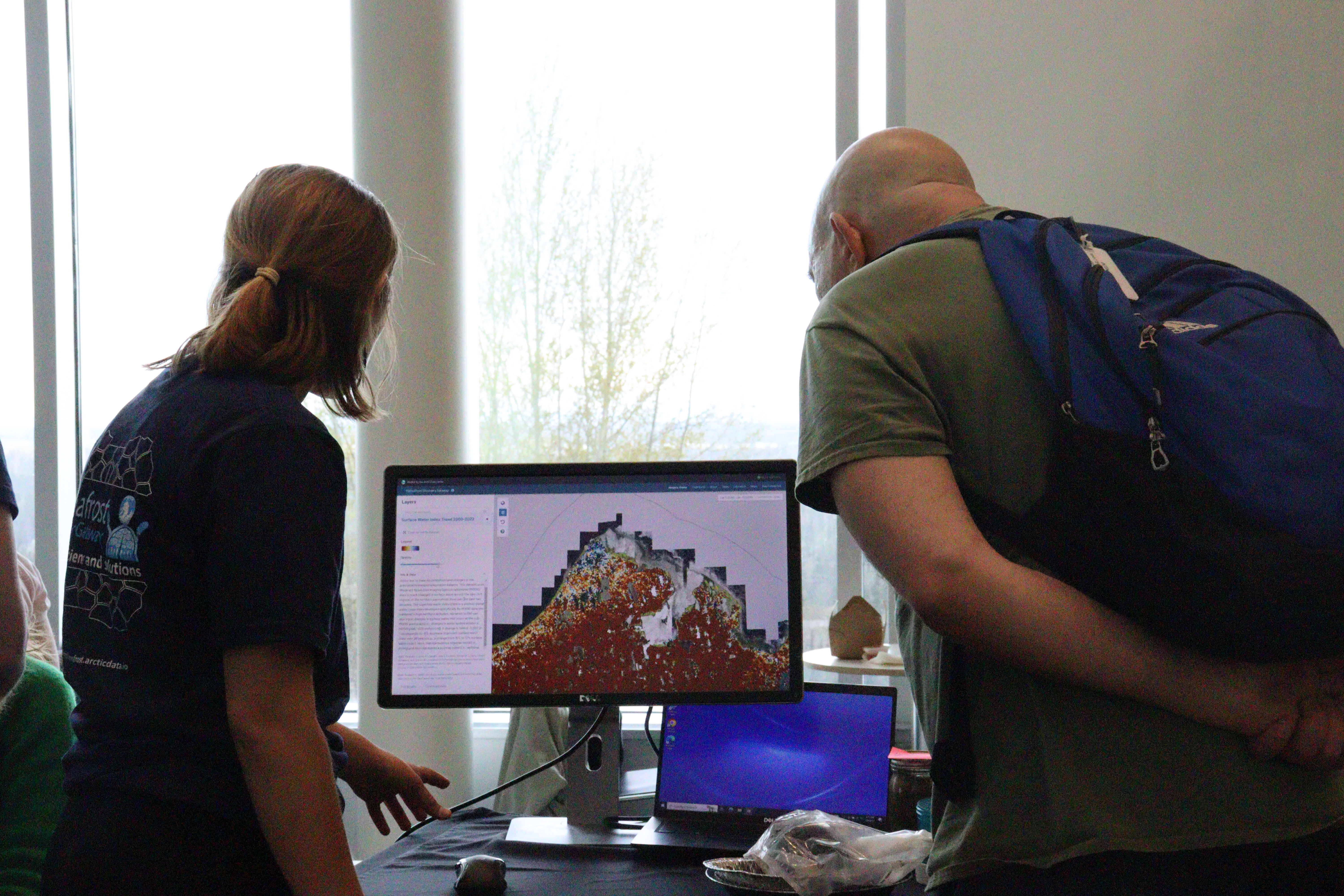 A woman, left, and a man, right, examine a computer monitor displaying the Permafrost Discovery Gateway interface, which looks like a map covered in different colored squares. 