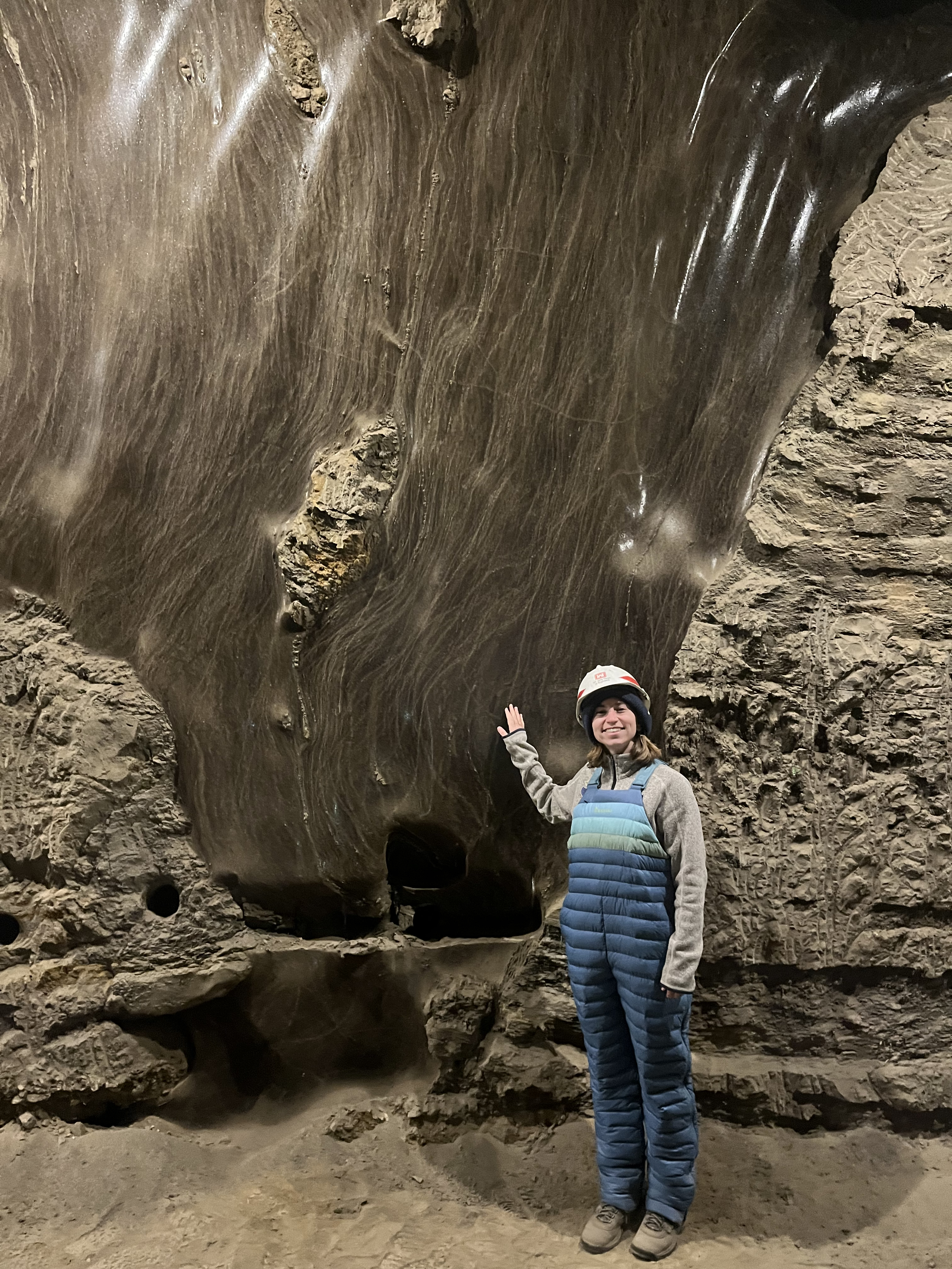 A woman stands next to a wall of soil, a vertical portion of which is darker and shinier than the rest. This portion is an ice wedge. 